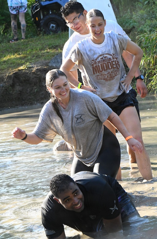 A group of smiling people in athletic gear run in a line into a ditch filled with muddy water.