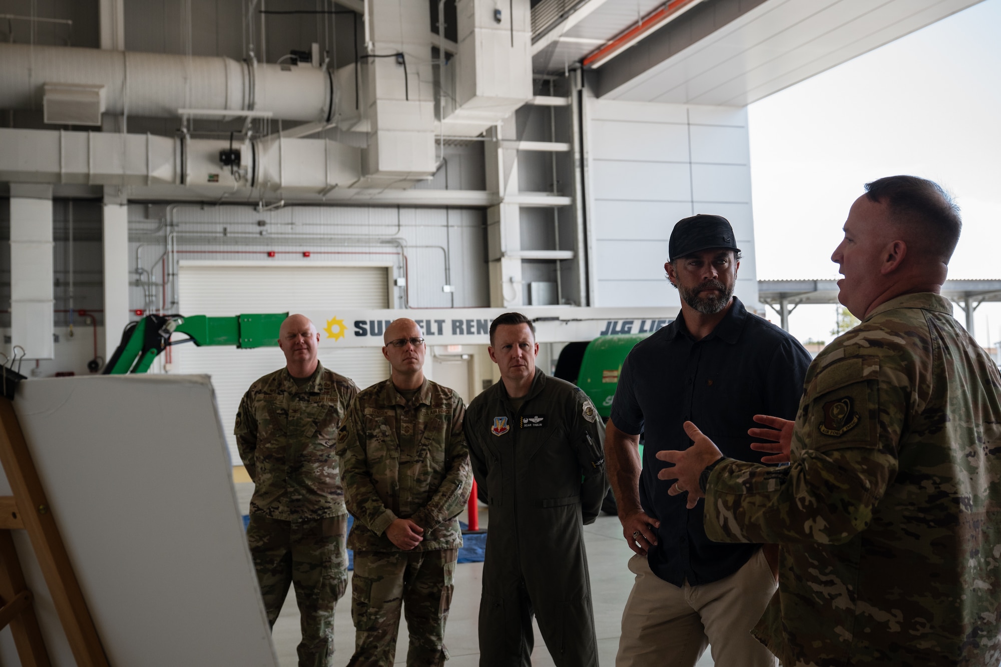 A man in a camouflage military uniform gestures as he speaks to a group of three other military members and one civilian in an aircraft hangar.