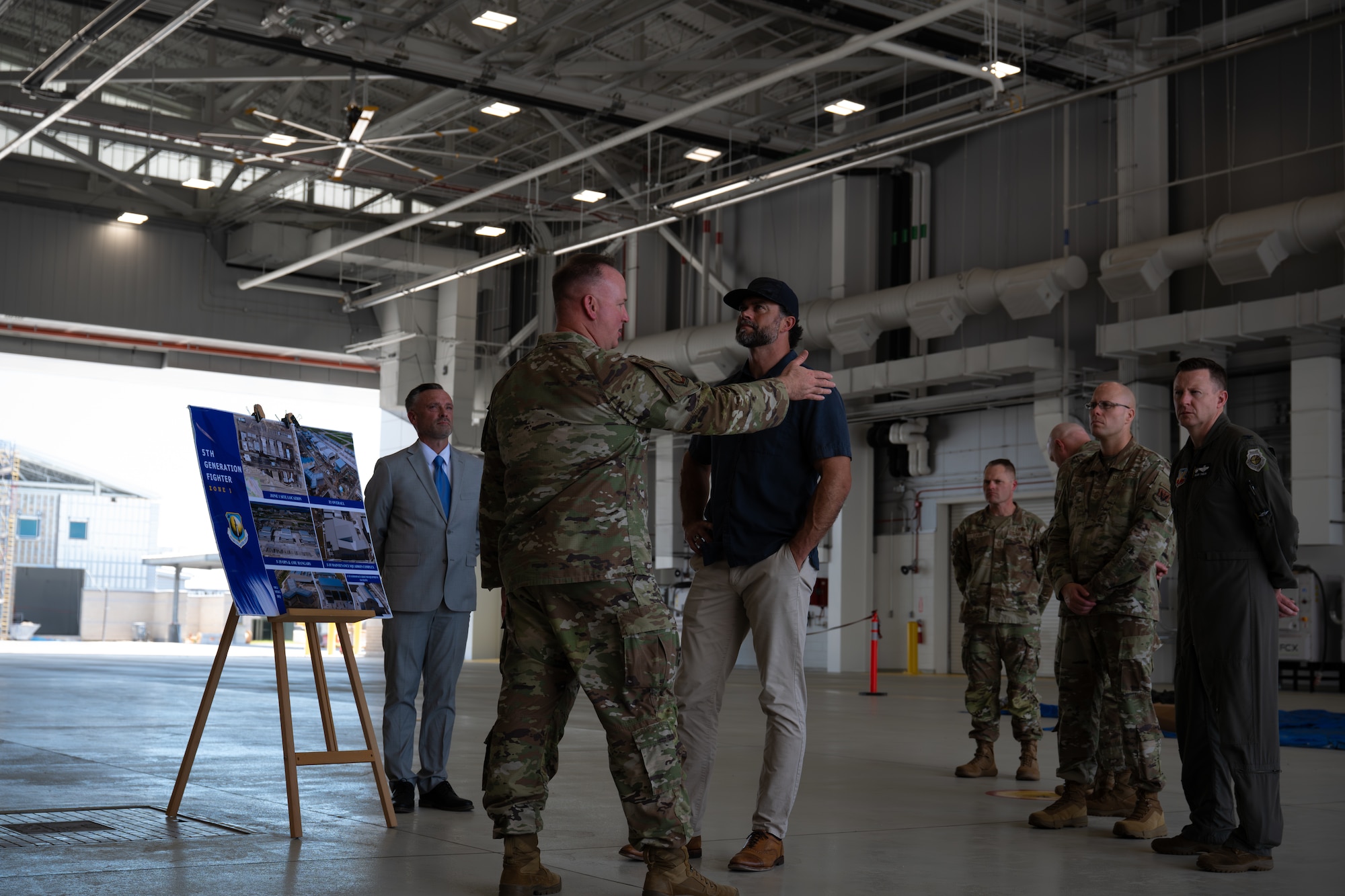 Inside an aircraft hangar, a man in a military uniform talks with a man in civilian clothes, placing a hand on his shoulder. In the background, other men look on near a presentation board.
