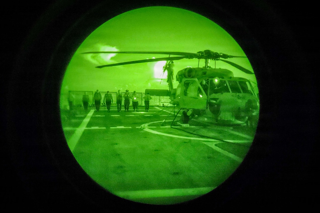 Sailors walk in a row near a helicopter on a ship's flight deck, seen through the green light of a night-vision lens.