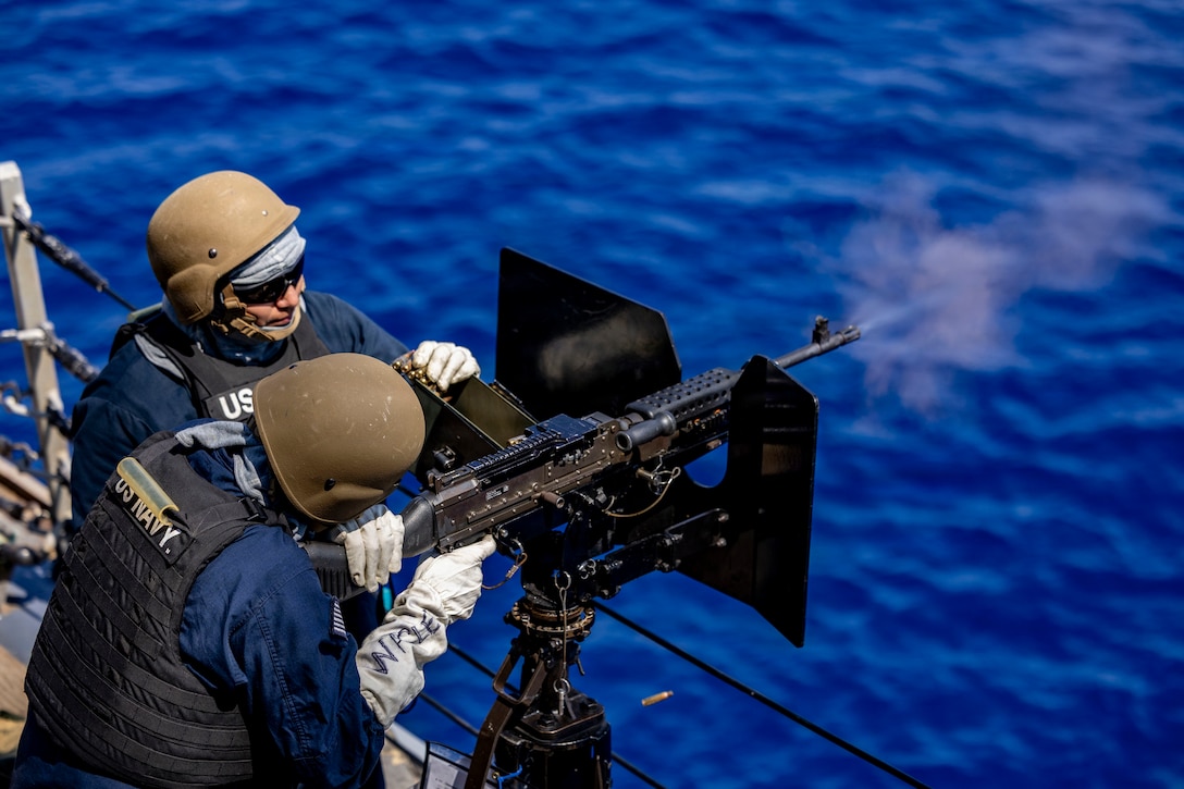 A sailor fires a mounted machine gun from a ship's deck over rich blue water as another sailor observes.