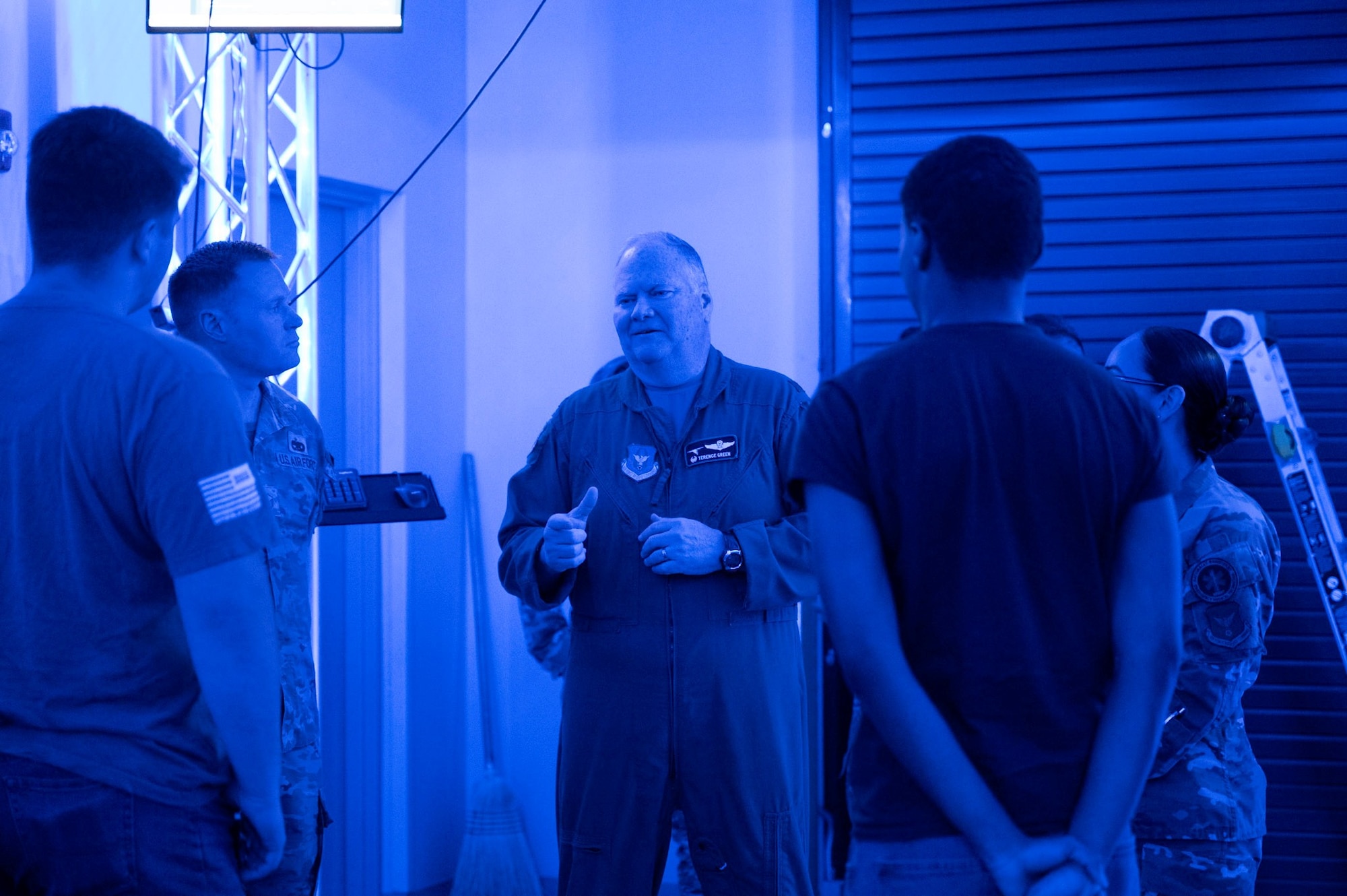 A man speaks to a group of people while standing in a room lit with blue lighting.