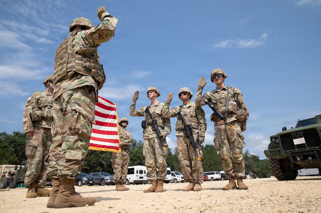 Three soldiers raise their right hands while facing a general doing the same as other troop hold a U.S. flag outdoors.
