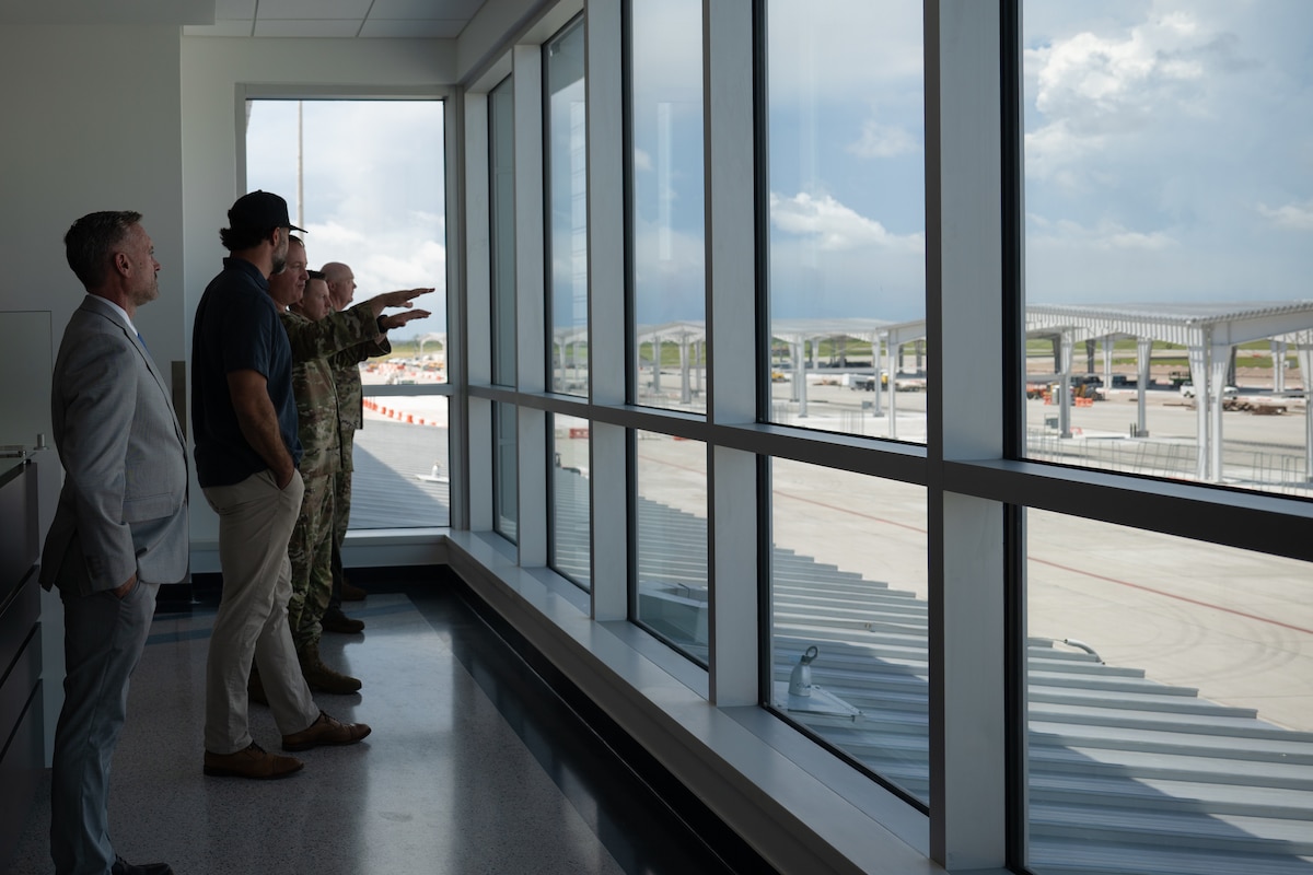 A man in a camouflage military uniform gestures as he speaks to a group of three other military members and one civilian in an aircraft hangar.