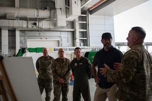 A man in a camouflage military uniform gestures as he speaks to a group of three other military members and one civilian in an aircraft hangar.