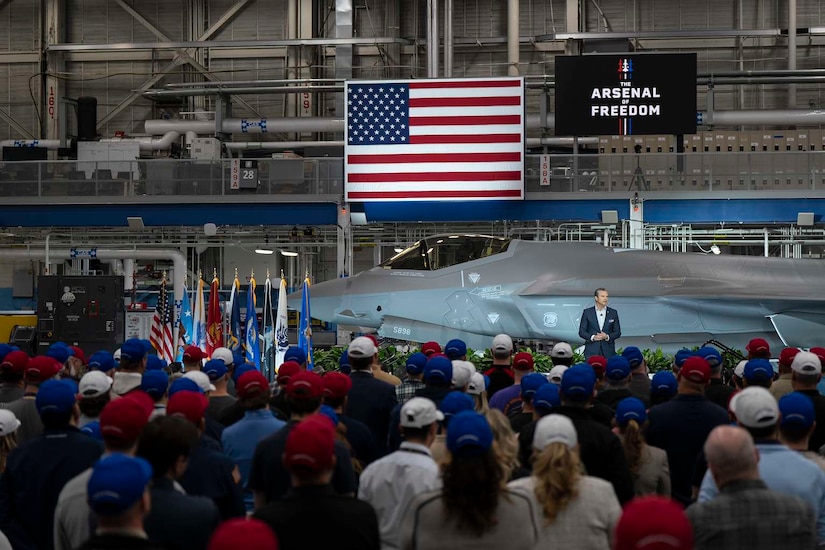 A man in business attire stands on a stage in a hangar and speaks to a large crowd of people in casual attire and baseball caps; behind him is a military aircraft, an American flag and a banner that reads, “Arsenal of Freedom.”