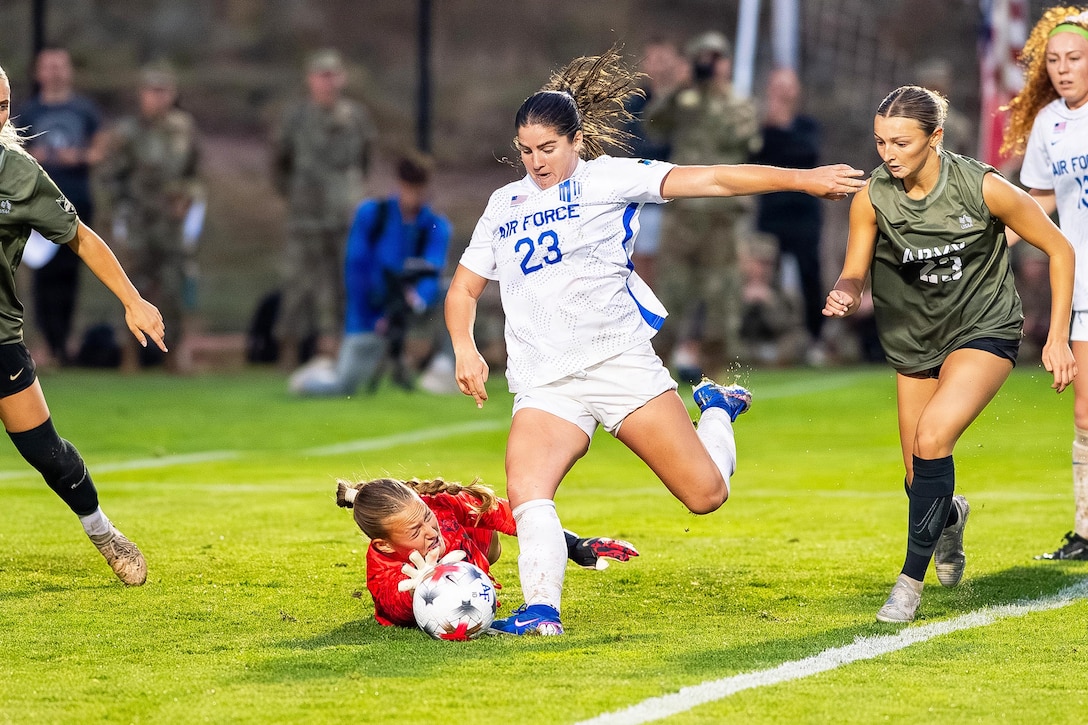 An Air Force soccer player attempts to kick the ball during a game as a goalie lying on the turf tries to block it.