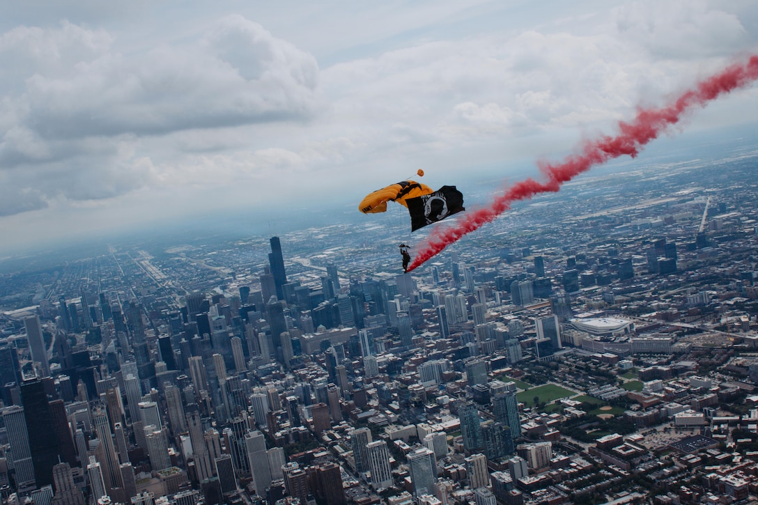 A soldier descends with a gold and black parachute and black flag over a city, leaving behind a red trail of smoke.