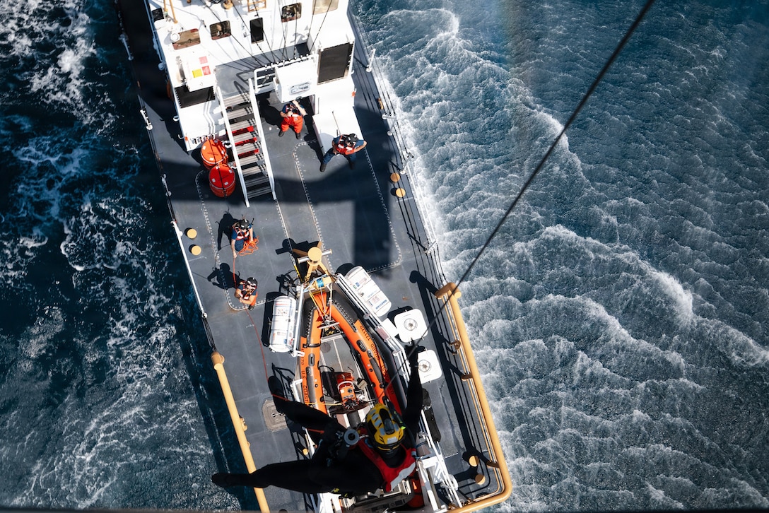 A Coast Guardsman is lowered with a line from a hovering helicopter to a ship at sea during the day.