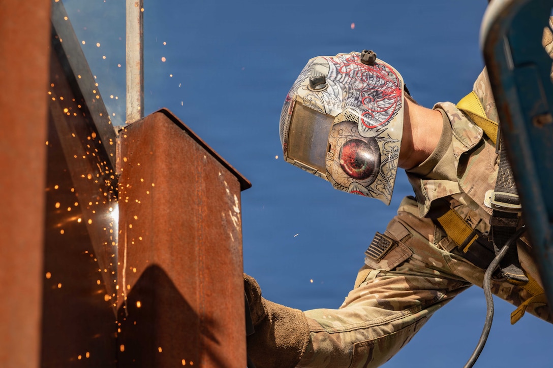 A close-up of sparks flying as a soldier wearing a protective face mask leans over a lift to weld parts of a fence against a blue sky.