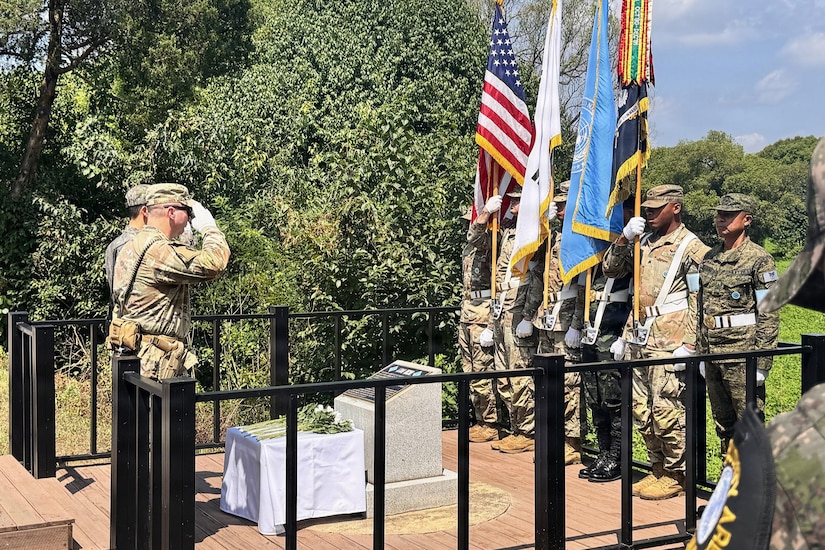 Two people wearing camouflage military uniforms stand in front of a memorial saluting as six other people in similar attire stand at attention holding several flags.