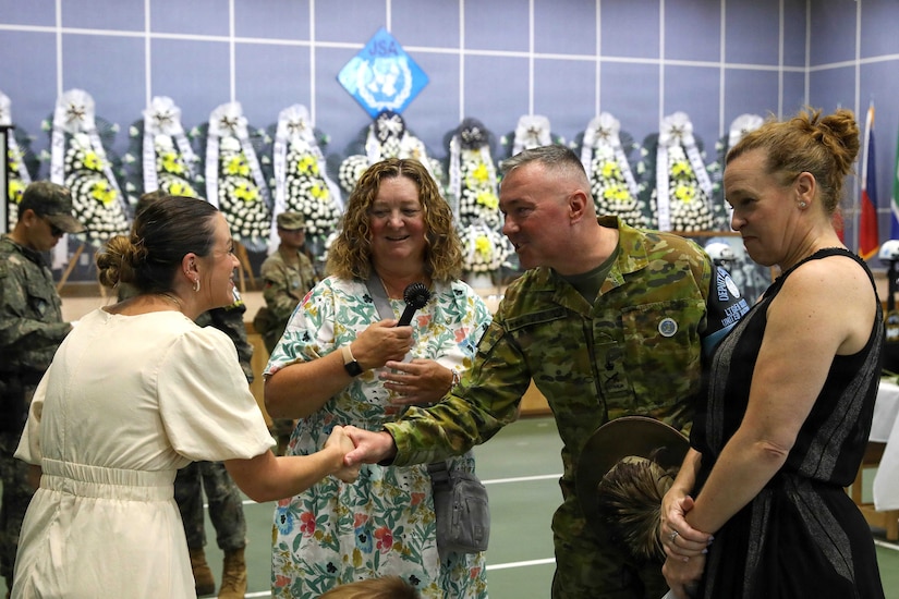 A man wearing a camouflage military uniform shakes the hand of a woman in formal attire while standing in an auditorium next to two other women in similar attire.