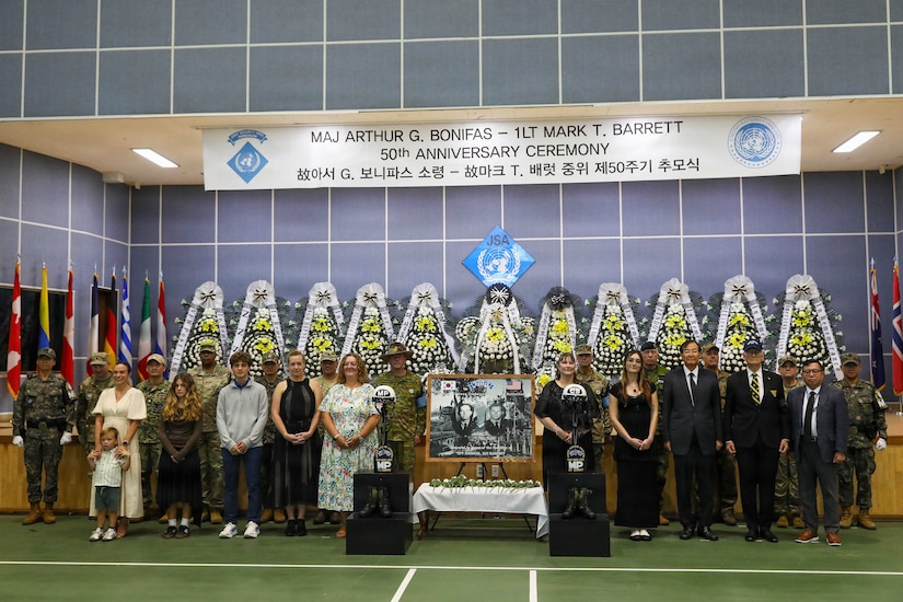 Approximately two dozen people wearing formal attire and camouflage military uniforms pose for a photo in a large auditorium while standing next to flower arrangements, a commemorative poster and two battlefield crosses.