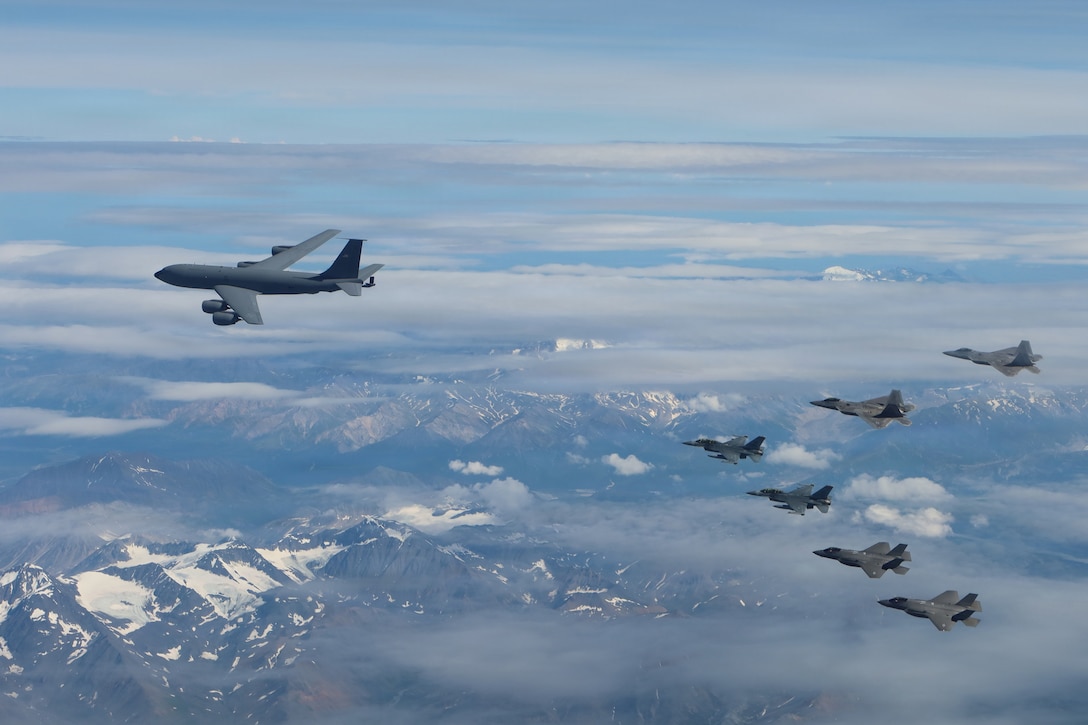 A large gray military jet aircraft flies followed by six smaller gray fighter jet planes in the air over snow-capped mountains during daytime.