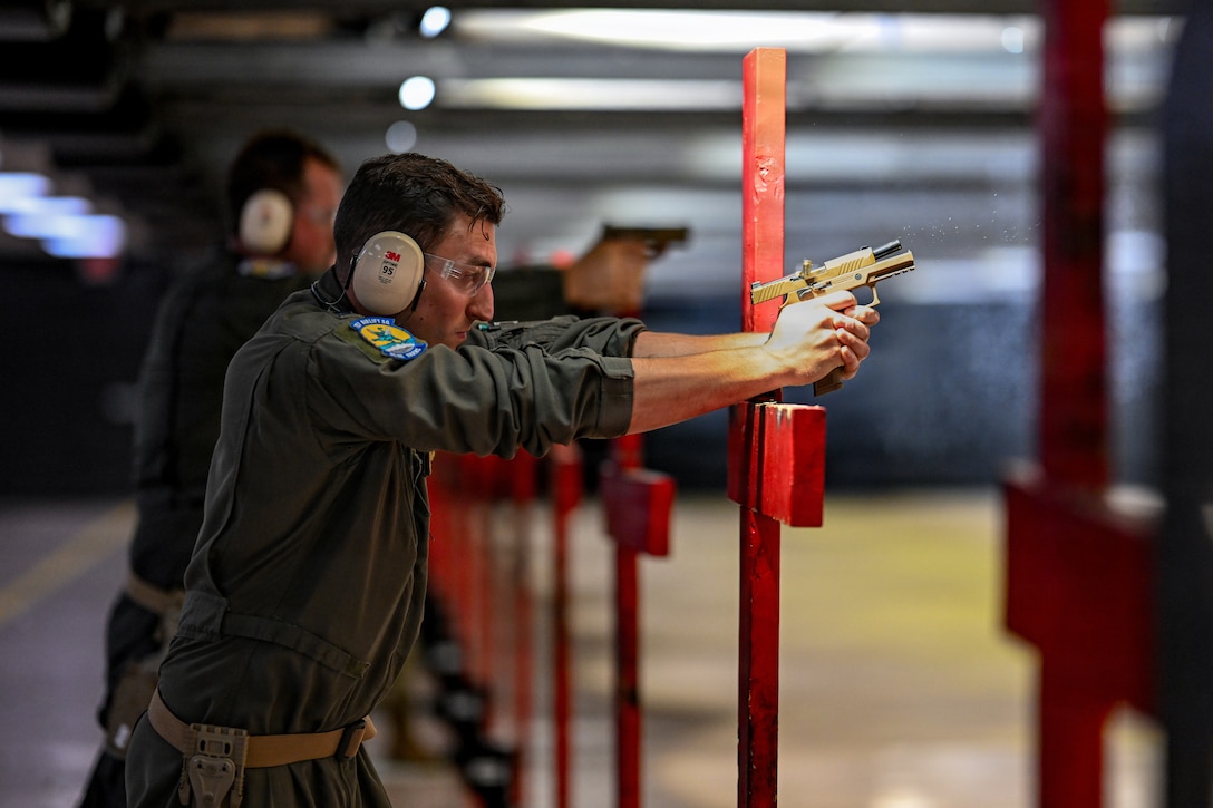 An airman wearing a flight suit, earmuffs and goggles fires a handgun on an indoor range, with another airman doing the same thing next to him.