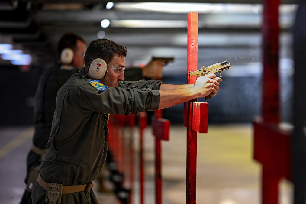 An airman wearing a flight suit, earmuffs and goggles fires a handgun on an indoor range, with another airman doing the same thing next to him.