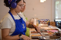 A woman wearing a blue apron and large blue headphones is stirring food in a large pan while another person watches nearby.