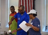 Three people wearing casual attire are standing near a small stove while the man in the middle stirs food in a frying pan. There is a US flag in the background.
