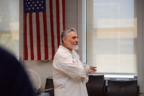Man in white chef's outfit is gesturing while talking, and in the background is a US flag on the wall.