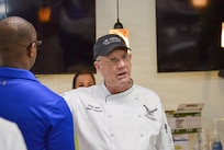 Man wearing white chef's uniform with dark blue baseball cap is gesturing while speaking to other people nearby.