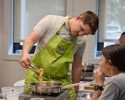A man in a light green apron is stirring food in a frying pan while someone else watches.