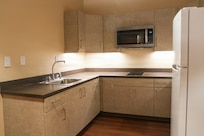 View of an empty kitchen with an L-shaped counter, sink, microwave and refrigerator.