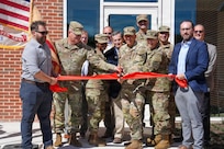 People wearing civilian attire and Army combat uniforms are participating in a ribbon-cutting for a new building.