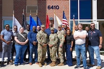 Fifteen people are standing in two rows posing for a picture in front of a building and various flags. The three people in the center are wearing Army combat uniforms, and they others are dressed in mixed attire.