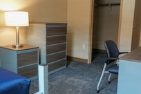 Empty bedroom is seen with a night stand, chest of drawers and desk with chair. There is an open door to a closet in the background.