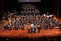 A large military band is performing on a stage with three members standing out in front of the band.
