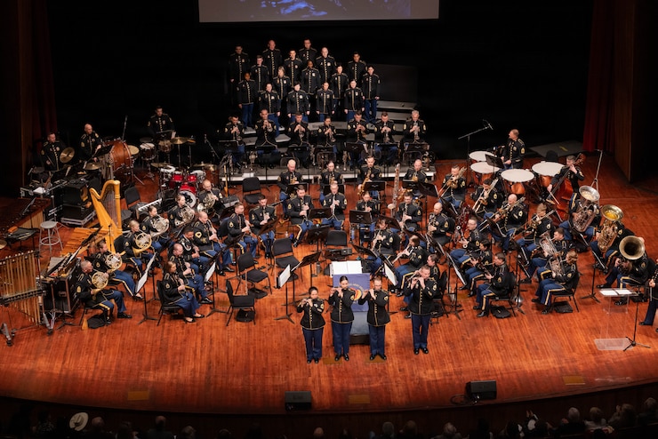 A large military band is performing on a stage with three members standing out in front of the band.