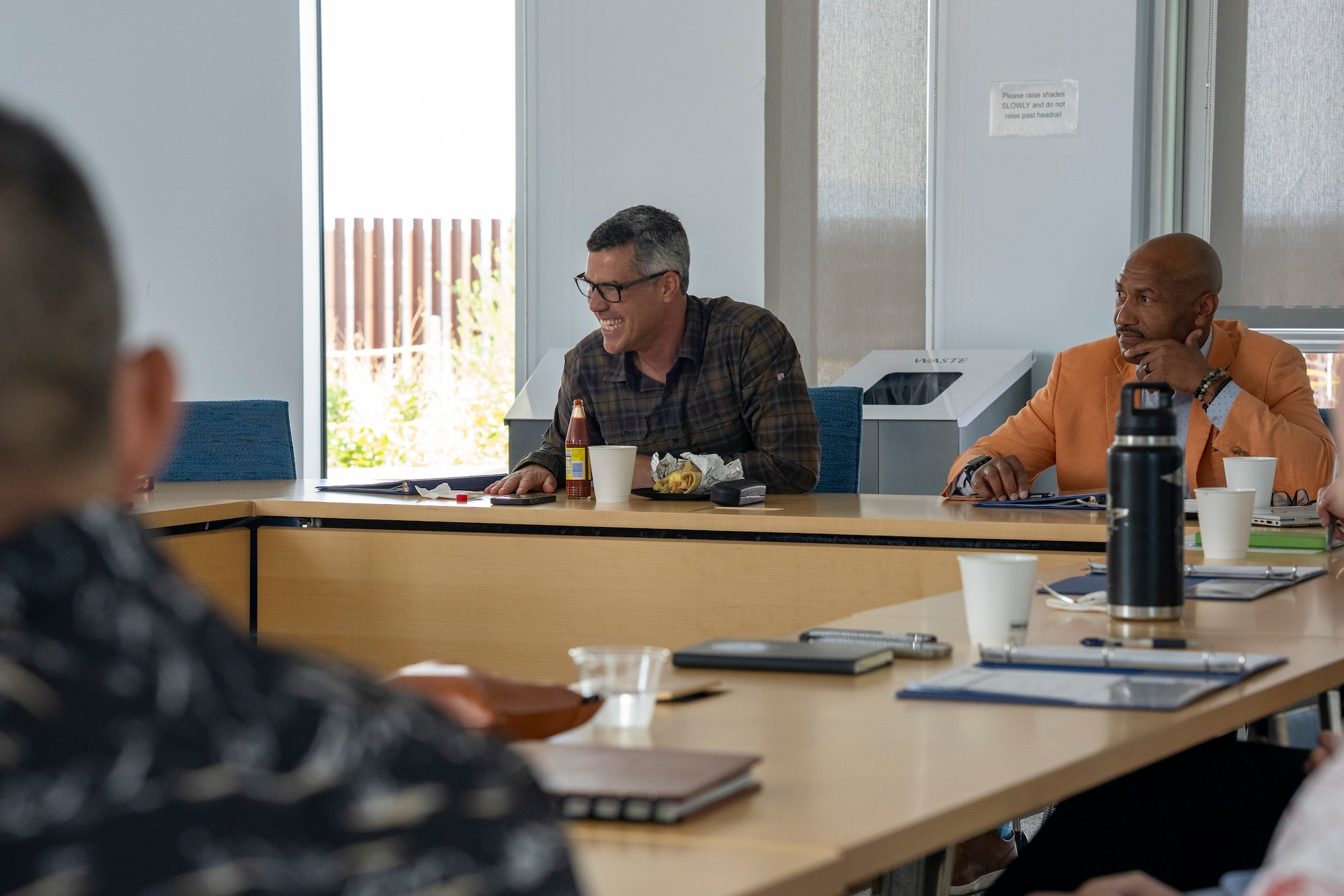 Two men sit at a table facing others. The man on the left is laughing.