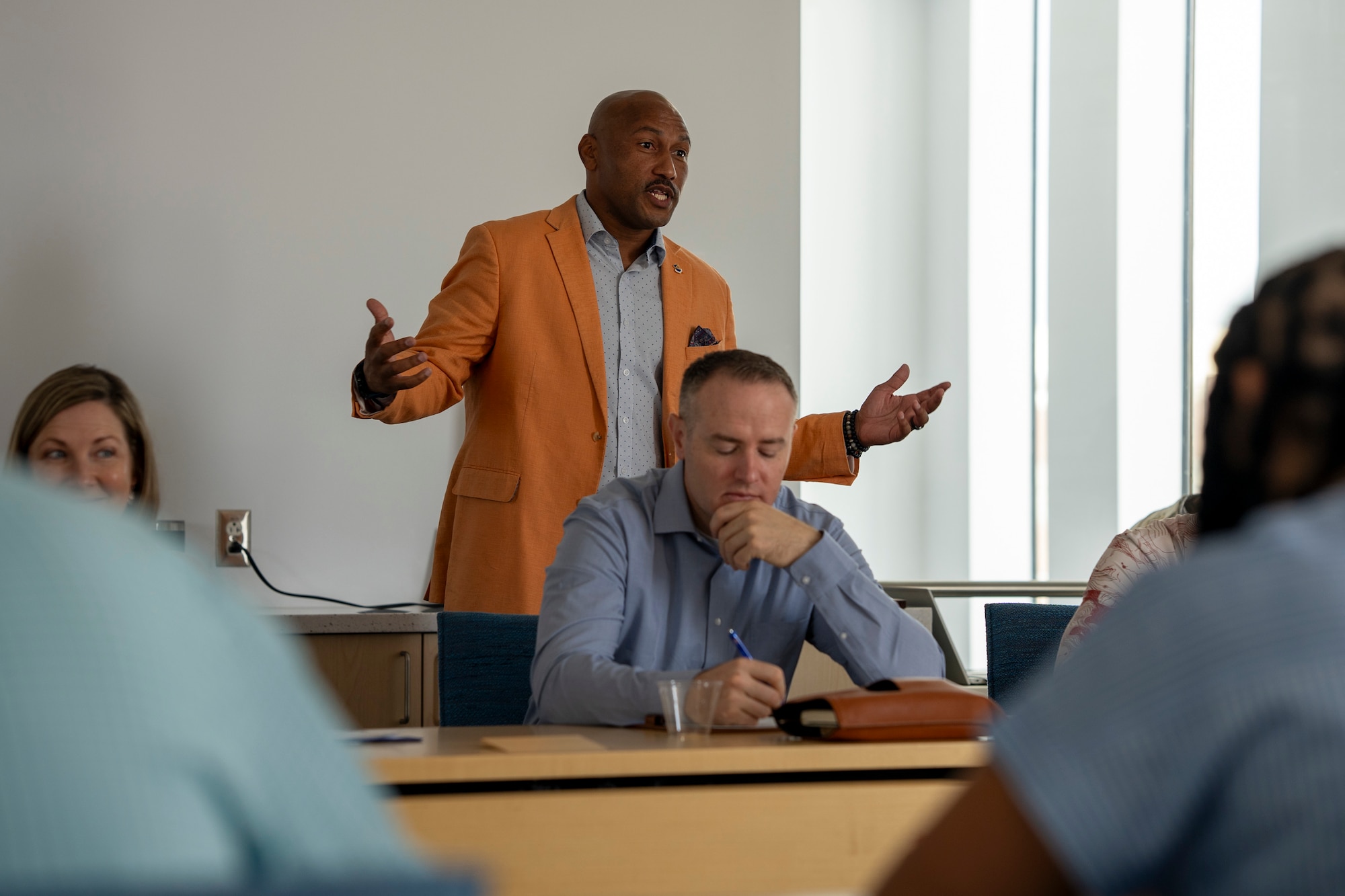 A man stands and spreads his arms as he addresses a group of people seated around a table.