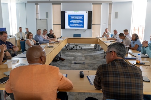 Primary focus is the back of two men who are facing a group of people seated around a large table.