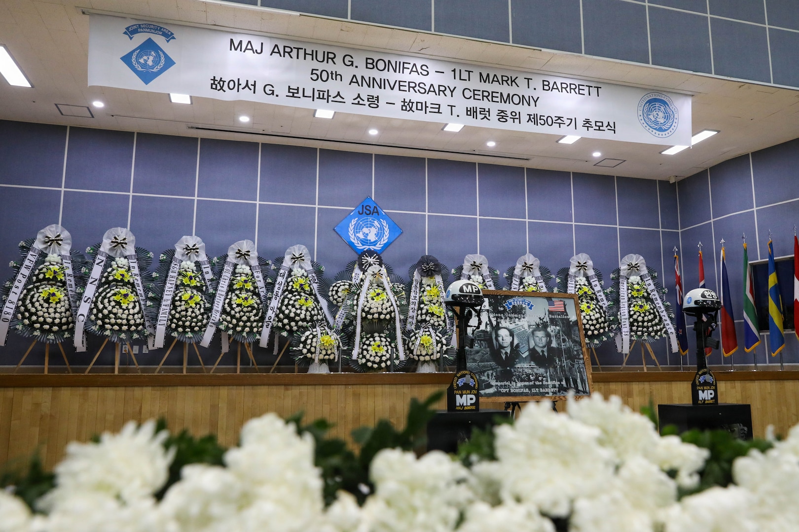 The 50th commemoration ceremony for Maj. Arthur G. Bonifas and 1st Lt. Mark T. Barrett takes place at Camp Bonifas on Aug. 18, 2026, with white chrysanthemums arranged in the foreground and the memorial stage visible in the background, honoring the two U.S. Army officers killed in the 1976 “axe murder incident” in the Korean Demilitarized Zone.