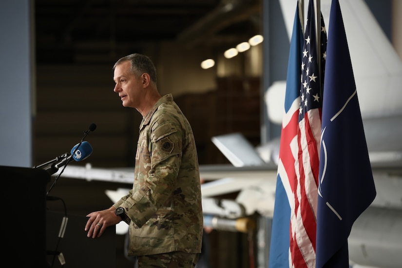 A man in a camouflage uniform speaks into a microphone while standing behind a lectern; the American, Iceland and NATO flags are displayed behind him.