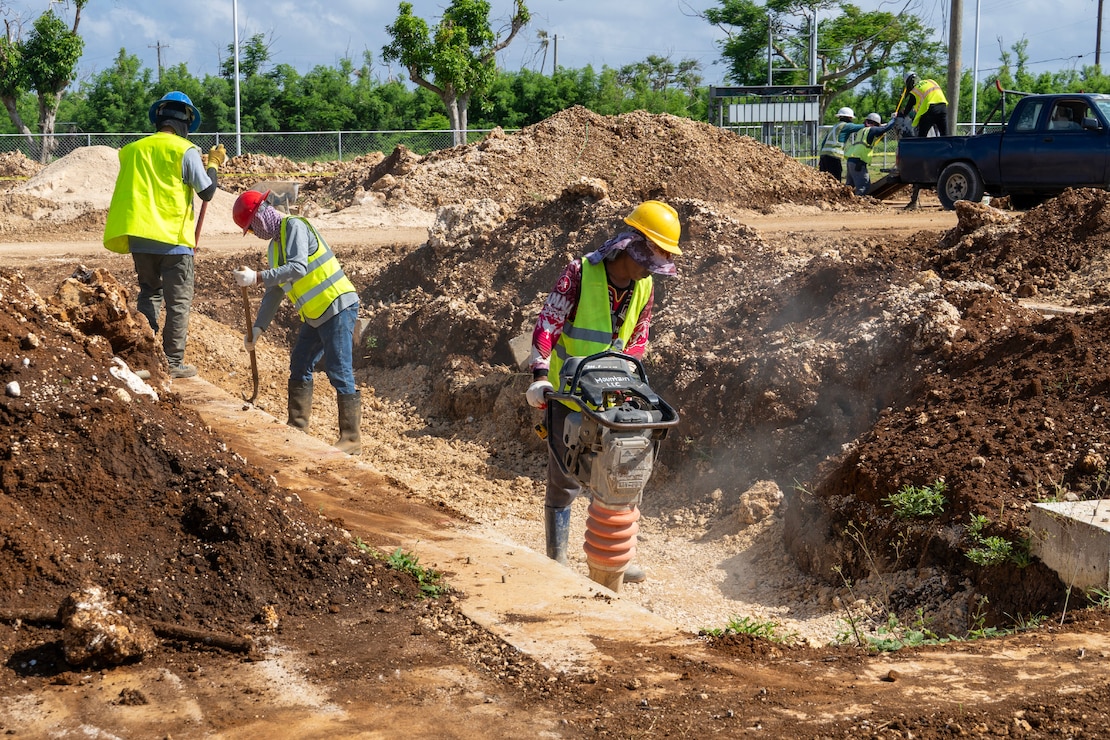 Construction workers wearing personal protective gear operate a device that tamps earth.