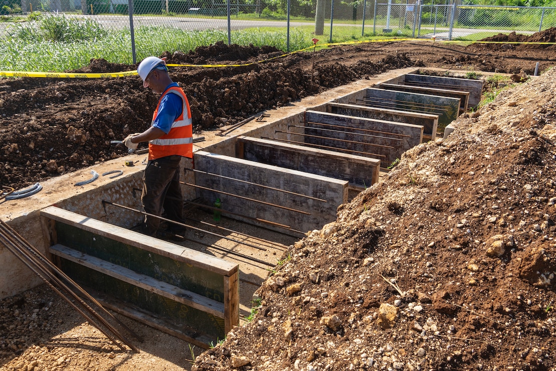 Steel rebar is connected to a concrete building foundation. A construction worker wearing personal protective equipment places a wood panel against the concrete foundation.