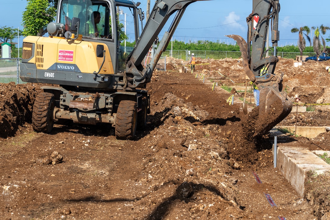 An excavator fills a trench next to concrete foundations with earth.