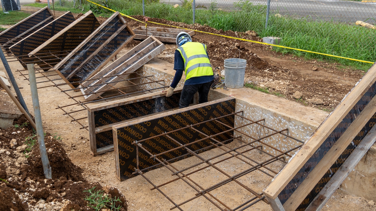 Steel rebar is connected to a concrete building foundation. A construction worker wearing personal protective equipment places a wood panel against the concrete foundation.