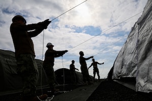 Five people in camouflage military uniforms and hard hats pull ropes to construct a tent outside.