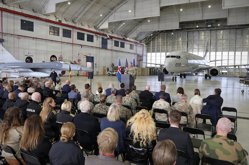A man in a camouflage military uniform speaks to dozens of people in similar attire and business attire seated in an aircraft hangar; there are two military aircraft in the background.