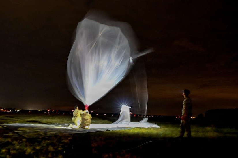 At night in an open field a person wearing a yellow safety suit holds onto a large balloon. Another person in a camouflage military uniform stands by to watch.