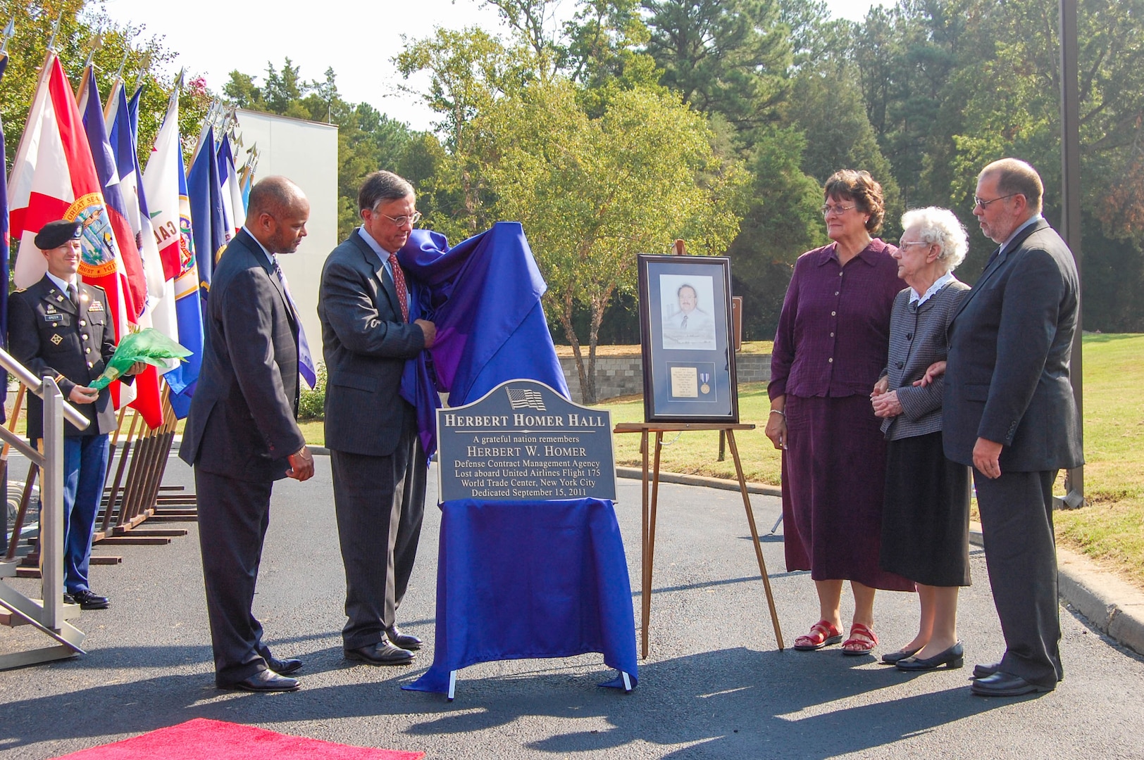 Four people watch as a person unveils a metal plaque at an outside event.