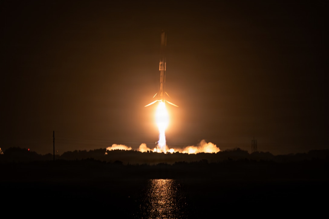 Flames erupt from a rocket taking off at night near a body of water.