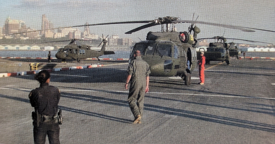 Two Army helicopters sit on a landing pad with a city skyline in the background.