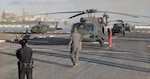Two Army helicopters sit on a landing pad with a city skyline in the background.