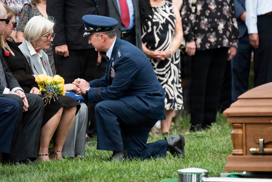 An airman in a dark blue military dress uniform kneels and holds hands with a seated woman near a coffin with other people nearby outdoors.