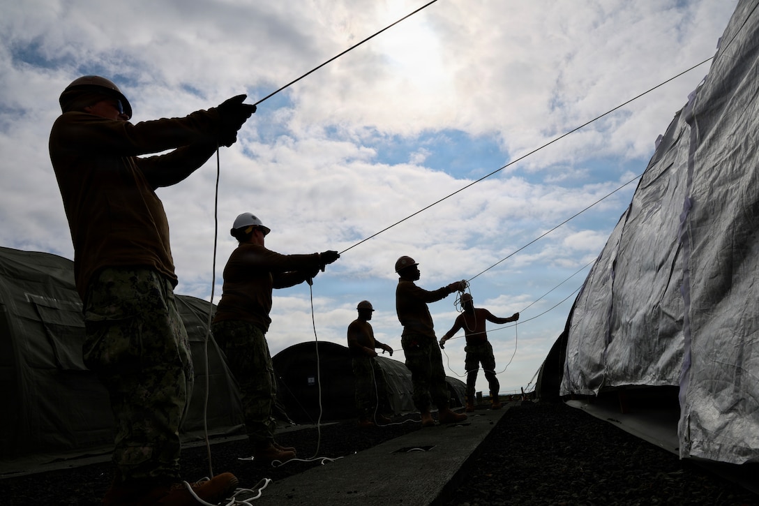 Five sailors wearing hard hats and shown in silhouette pull ropes to erect a large tent outdoors during daytime.