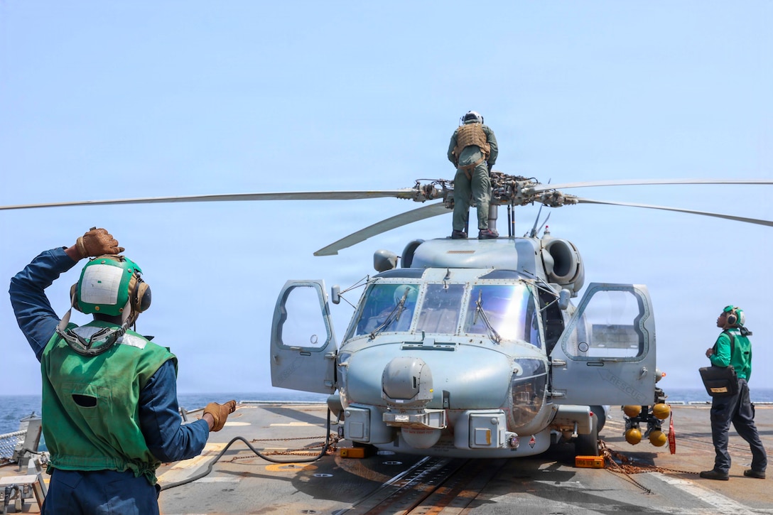A sailor in a green jumpsuit stands on top of a helicopter parked on a ship's deck and looks at the rotor as two other sailors on deck watch.